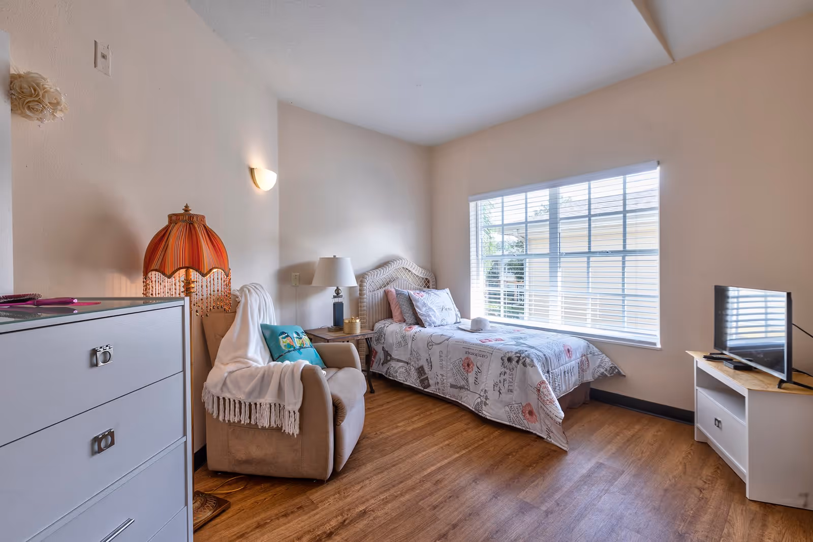 A cozy bedroom with a single bed covered in a patterned bedspread, a beige armchair draped with a white blanket and a decorative pillow, a wooden nightstand with a lamp, a dresser, and a TV on a stand near a large window with blinds.