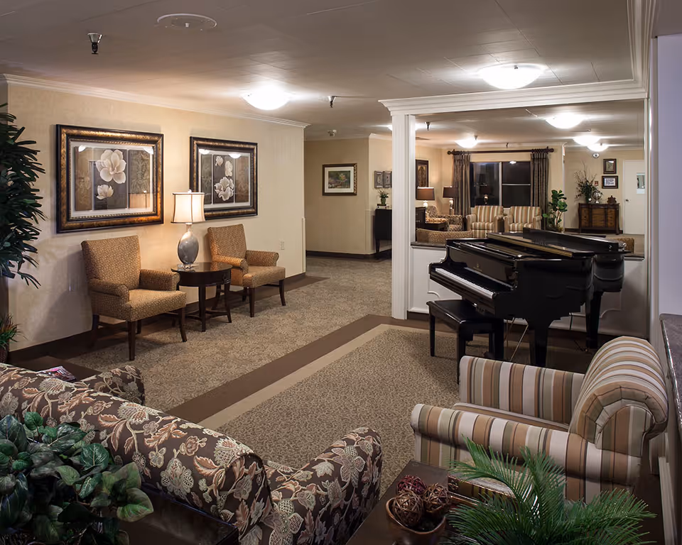 A cozy living room area in a senior living facility featuring a grand piano, several upholstered chairs and sofas with floral and striped patterns, framed floral artwork on the walls, table lamps, and decorative plants. The room has warm lighting and a carpeted floor.