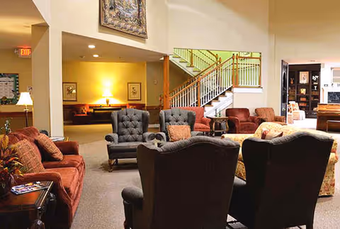 A spacious living room area in a senior living facility with multiple upholstered armchairs and sofas arranged around a central space. There is a staircase with a wooden railing in the background, warm lighting, and framed artwork on the walls.