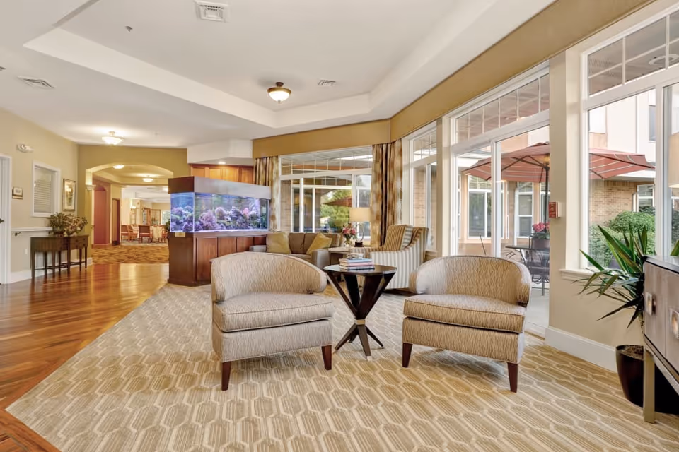 Bright senior living lounge with two upholstered chairs and a small table, a large aquarium, and floor-to-ceiling windows overlooking an outdoor patio.