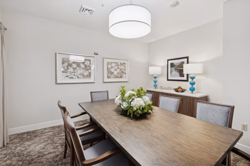 A dining room with a wooden table surrounded by six chairs. A floral centerpiece decorates the middle of the table. On the back wall, there is a wooden sideboard with two blue lamps and a framed picture above it. Two framed artworks hang on the adjacent wall. The room has a neutral color palette with beige walls and carpeted floor.