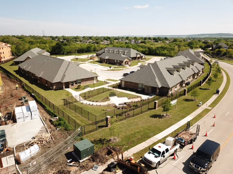 Aerial view of a senior living campus with multiple single-story brick buildings around a central courtyard, fenced lawns, and vehicles along the adjacent road.