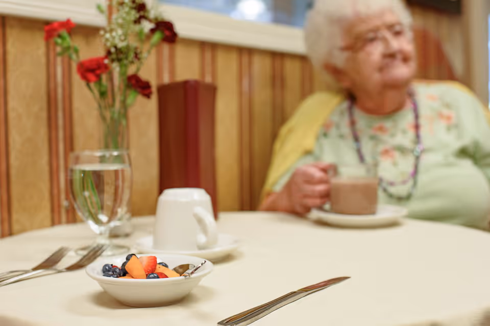 A small bowl of mixed fresh fruit on a table set with utensils, a glass of water, and a cup. In the background, an elderly woman wearing glasses and a floral shirt with a yellow cardigan is sitting and holding a cup, with a vase of red flowers on the table behind her.