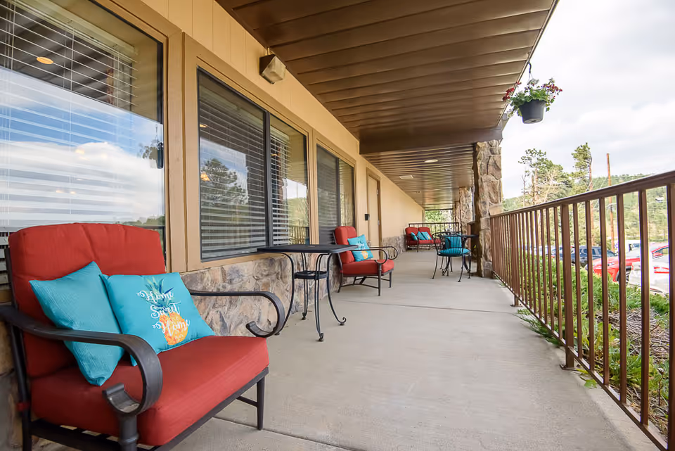 Covered outdoor patio area with red cushioned chairs and small black metal tables. The patio has a railing on one side and windows with blinds on the other. There are decorative pillows on the chairs and hanging flower pots above.