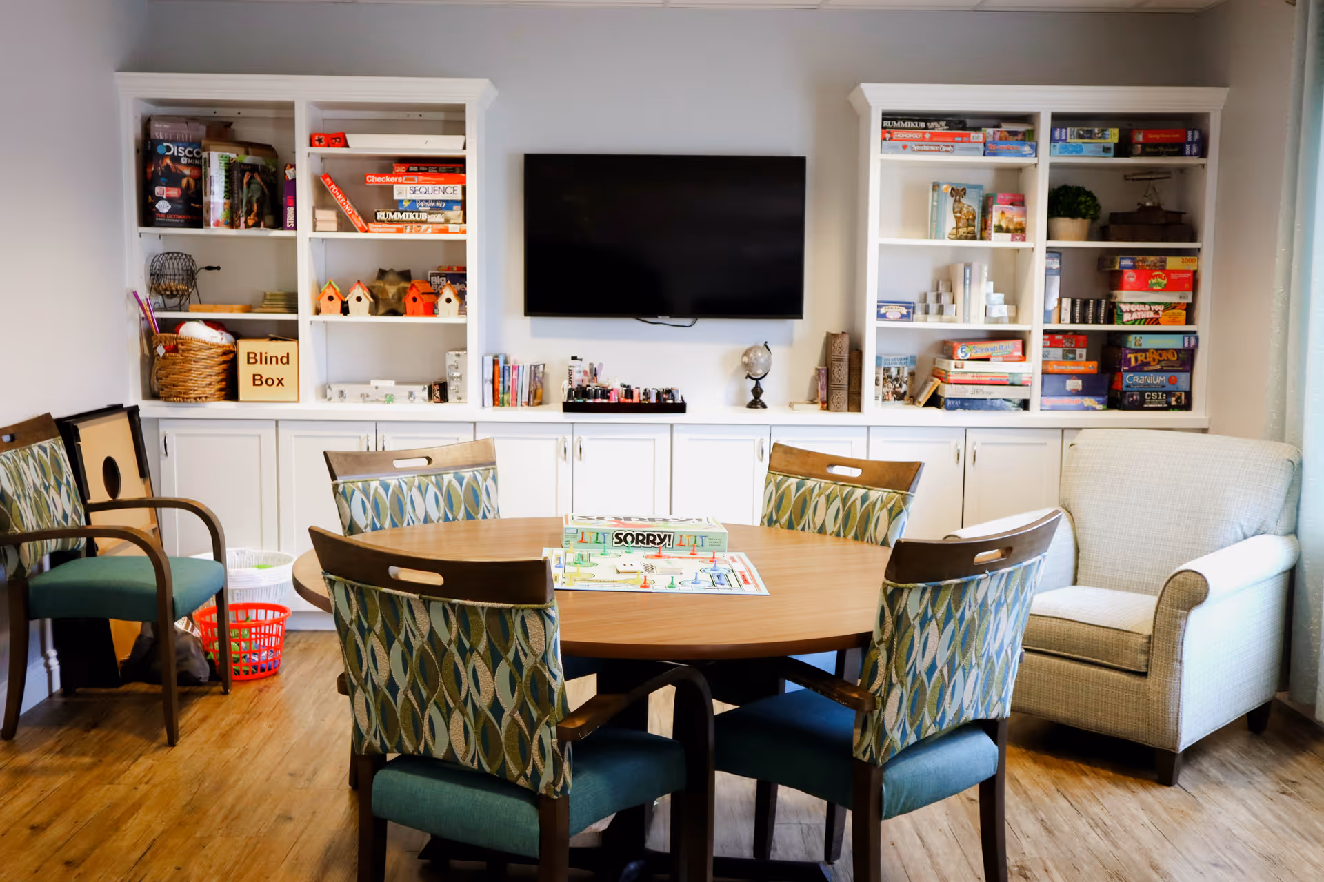 A cozy game room with a round wooden table surrounded by five patterned chairs. On the table, there are board games including Sorry! and Life. Behind the table, there are white built-in shelves filled with various board games, books, and decorative items. A flat-screen TV is mounted on the wall above the shelves. To the right, there is a comfortable upholstered armchair. The room has wooden flooring and light-colored walls.