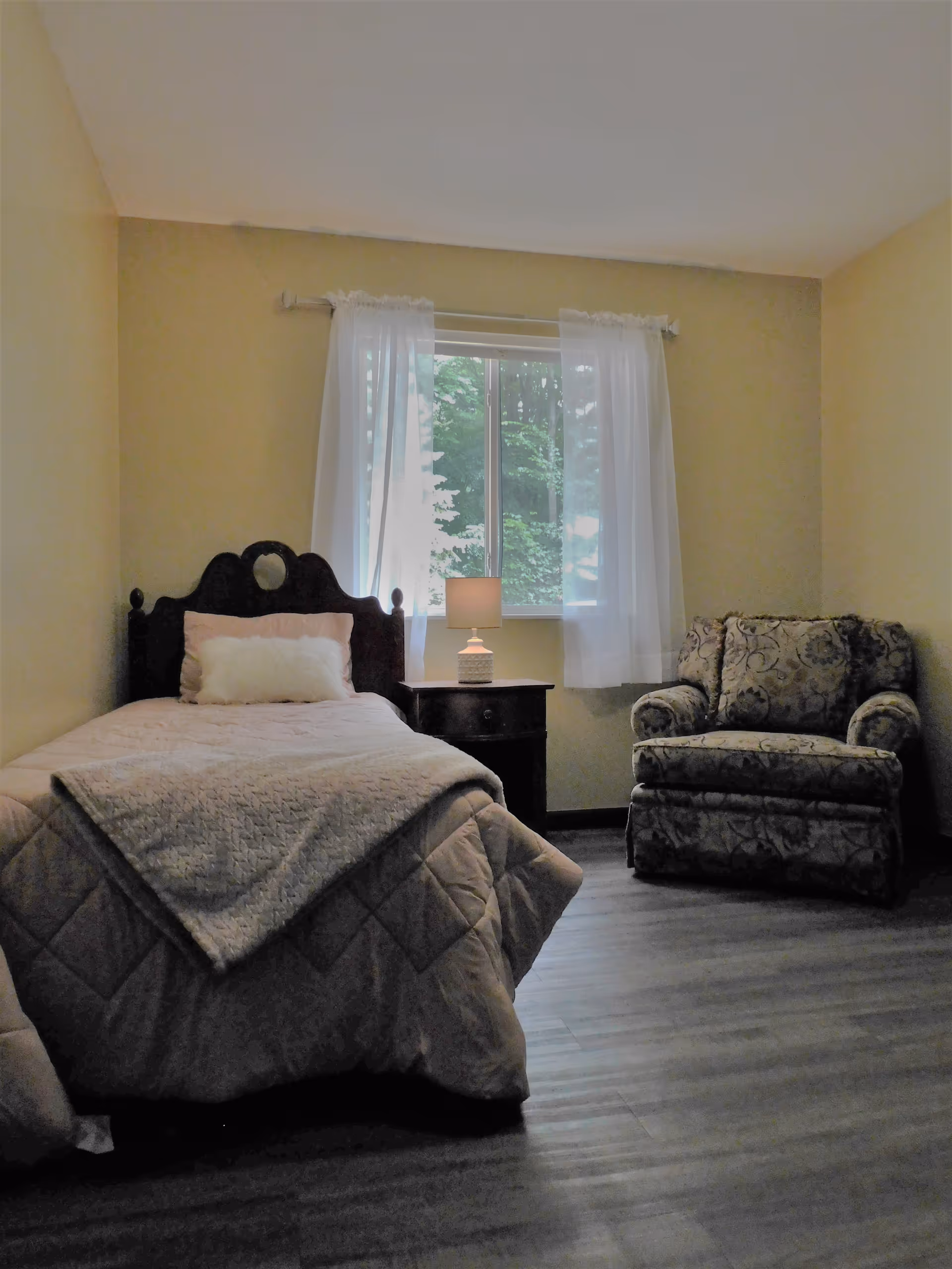 A cozy bedroom with a single bed covered in a light pink quilt and a fluffy white pillow. Next to the bed is a wooden nightstand with a small lamp. A window with sheer white curtains lets in natural light, and there is a patterned armchair in the corner of the room. The walls are painted a soft yellow and the floor is wood.