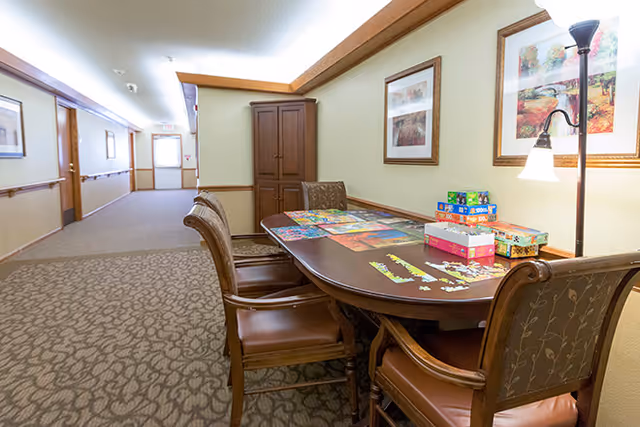 A hallway in a senior living facility with a table and chairs set up for puzzle and board game activities. The table has several puzzles and game boxes on it. The walls are decorated with framed artwork and there is a floor lamp providing light.