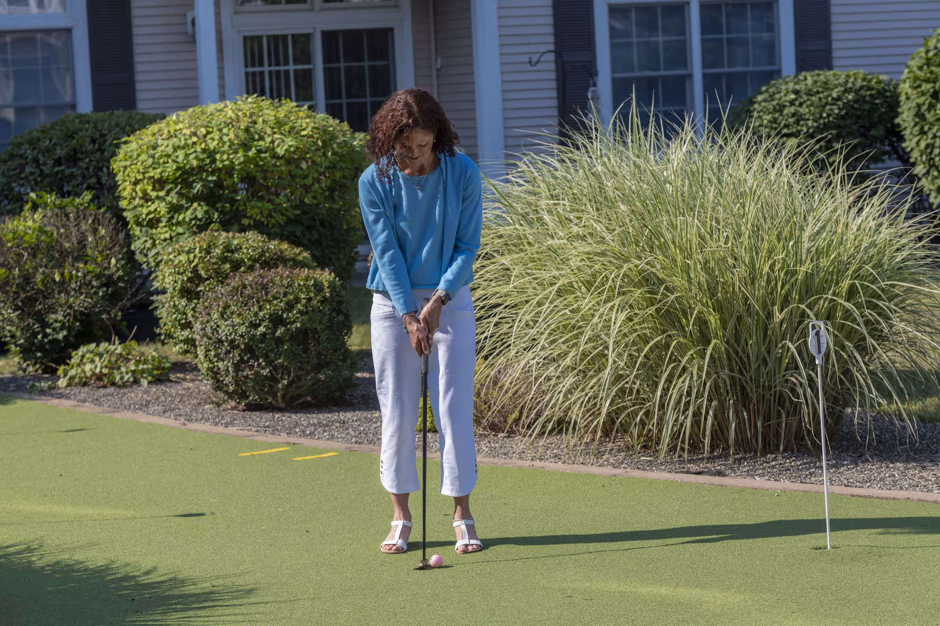 A woman wearing a blue long-sleeve shirt and white pants is playing mini golf on a green putting surface outside, with bushes and a building with windows in the background.