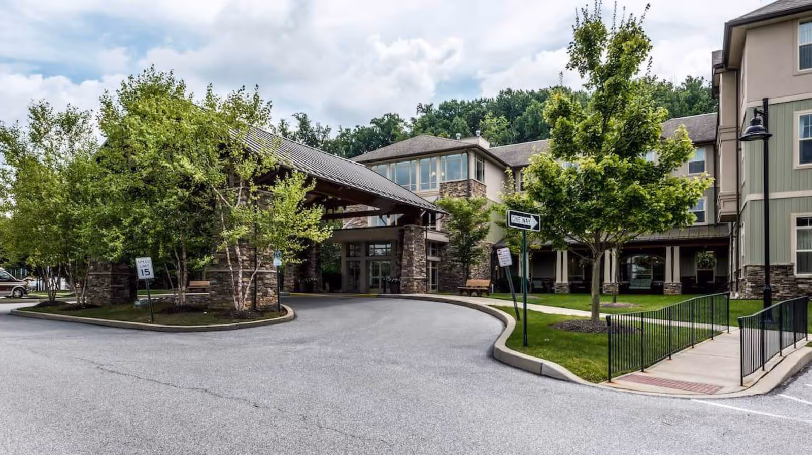 Front entrance of a multi-story senior living facility with a covered porte-cochere, driveway, trees, and signage.