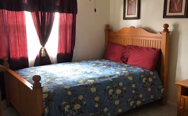 A bedroom with a wooden bed frame, a floral patterned blue bedspread, and two red pillows. There are red curtains covering a window behind the bed and two framed pictures hanging on the wall above the headboard.