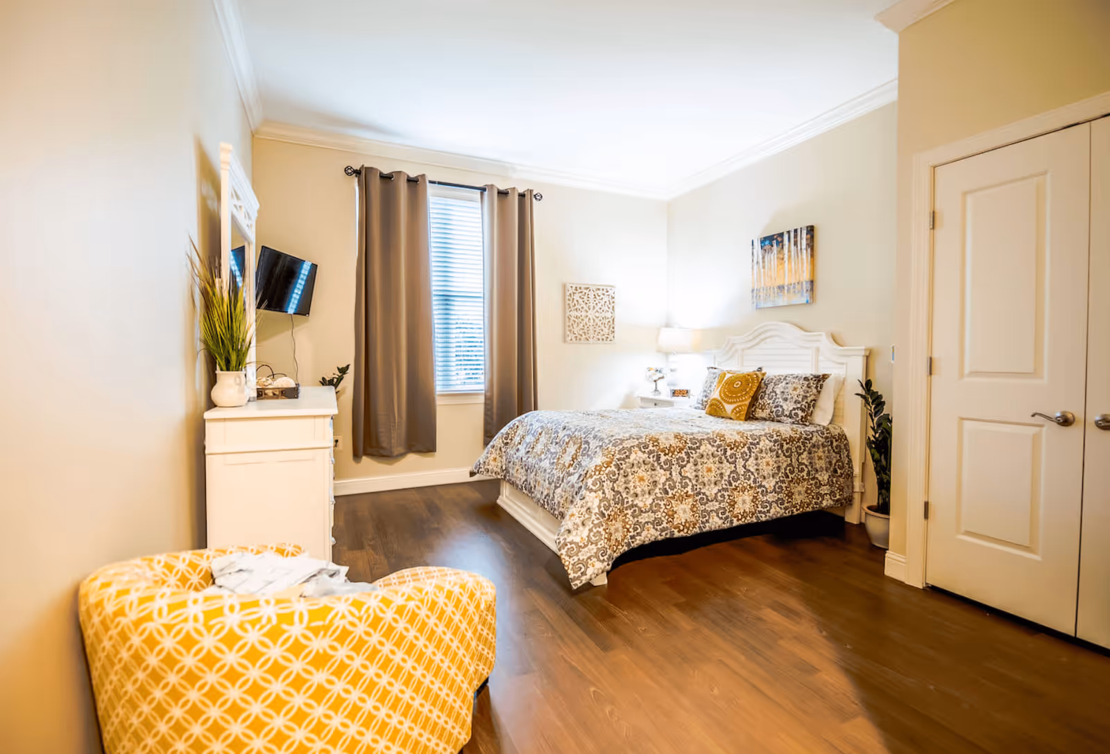 A cozy bedroom with a patterned bedspread on a white bed frame, a yellow patterned armchair, a white dresser with a potted plant and a wall-mounted TV, beige walls, a window with brown curtains, and wooden flooring.
