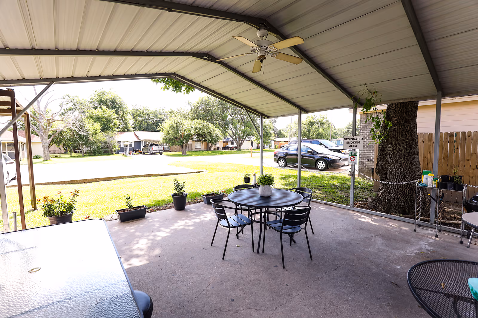 Covered outdoor patio area with a round black metal table and four matching chairs. Several potted plants are placed along the edge of the patio. A ceiling fan is mounted on the roof. In the background, there is a grassy lawn with trees, parked cars, and residential houses. A sign indicating a designated smoking area is visible near a large tree.