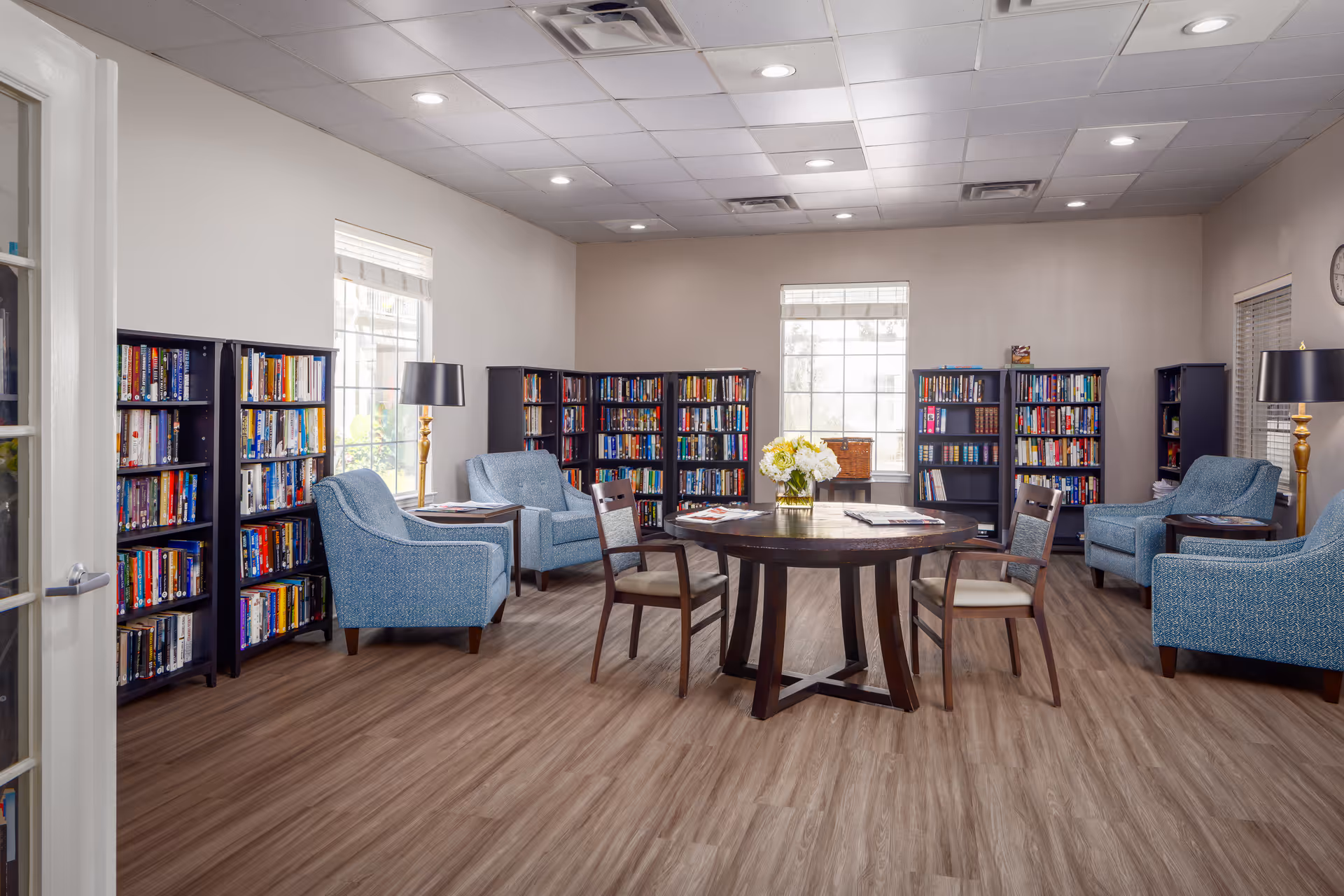 A bright and spacious reading room with multiple bookshelves filled with books along the walls. The room features four blue upholstered armchairs paired with small side tables, and a round wooden table in the center with four wooden chairs around it. A vase with flowers is placed on the round table. Large windows allow natural light to fill the room, and the floor is covered with wood-style flooring.
