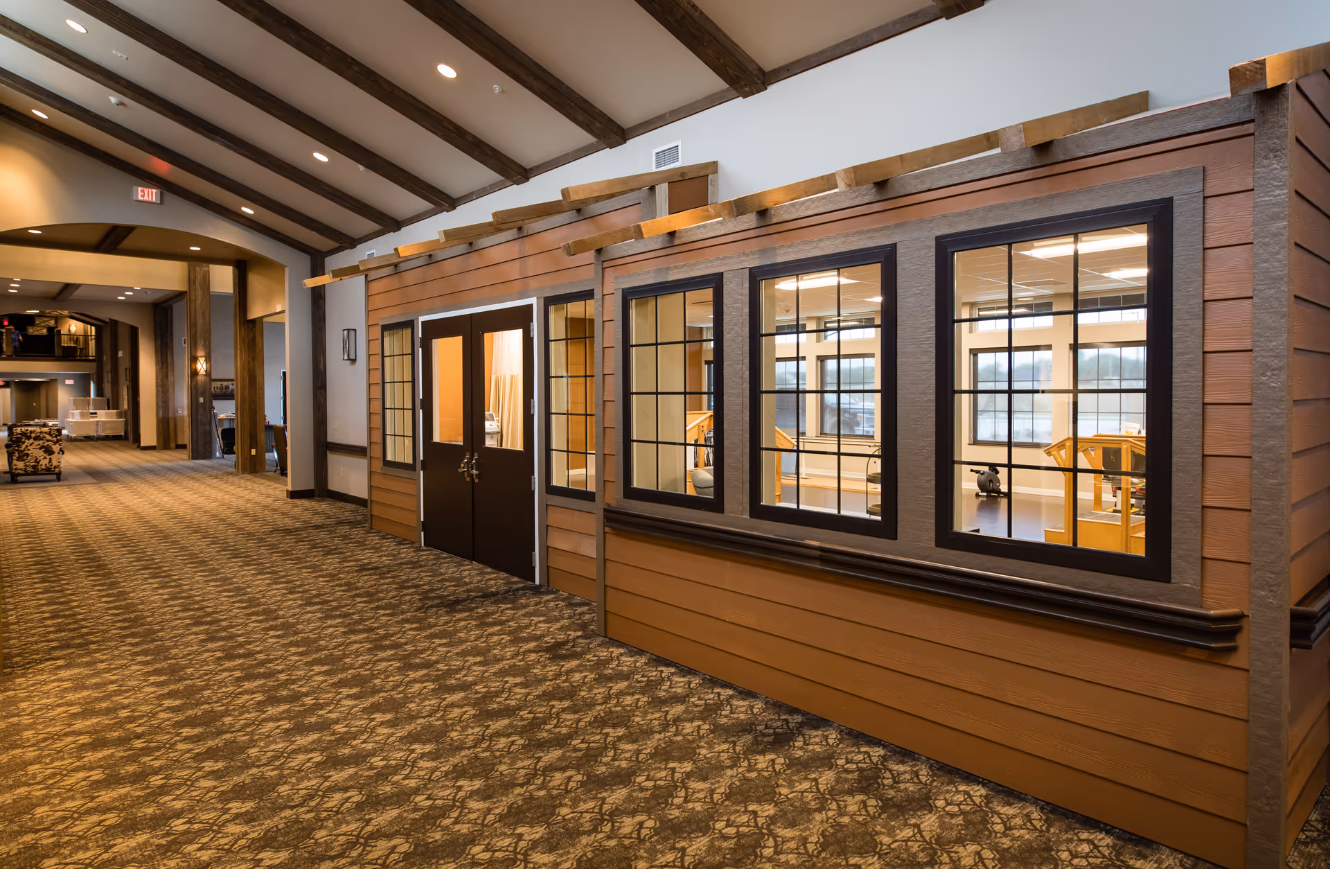 Interior hallway of a senior living facility with a carpeted floor and wooden beams on the ceiling. On the right side, there is a room with large windows and double doors, designed to look like the exterior of a house with brown siding and black window frames. Inside the room, exercise equipment and wooden furniture are visible. The hallway extends into the distance with additional seating and lighting.