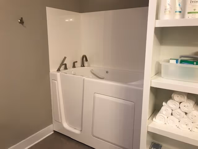 A walk-in bathtub with a door and safety grab bar, adjacent to a shelving unit stocked with rolled white towels and various toiletries.