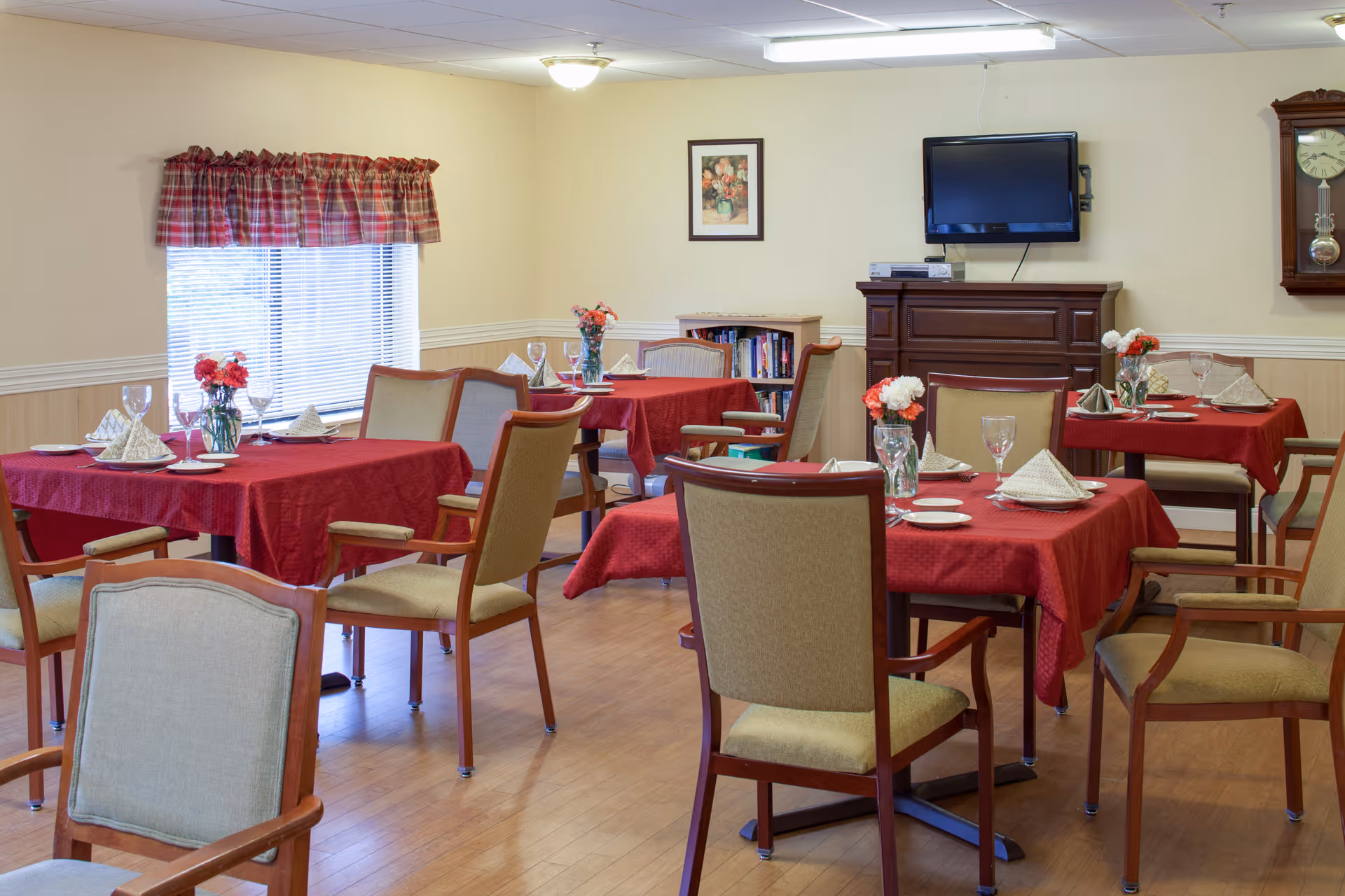 Dining room with several tables covered in red tablecloths set with glassware and napkins, wooden chairs, floral centerpieces, and a wall-mounted TV.