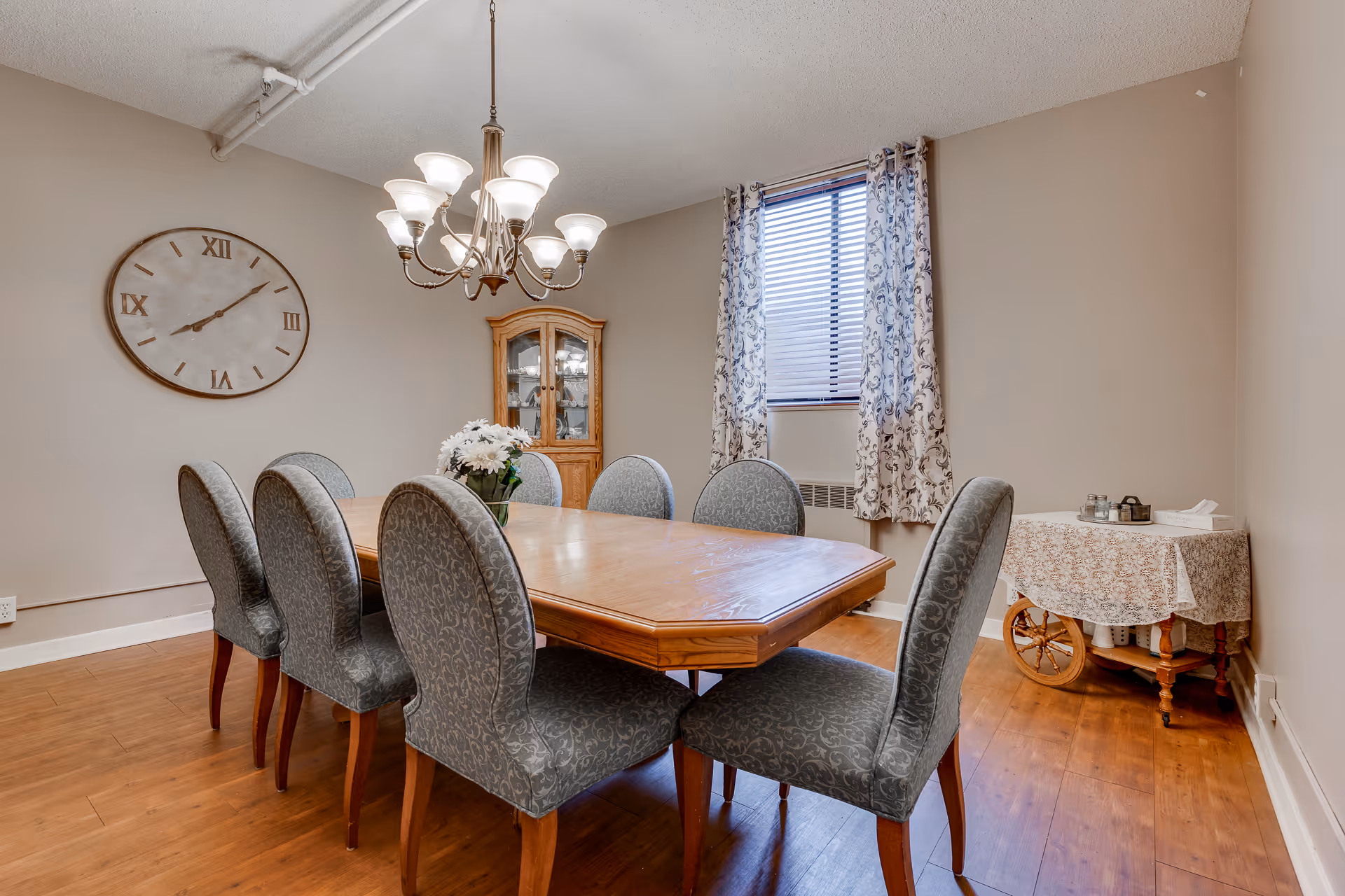 A dining room with a wooden table surrounded by eight upholstered chairs. A chandelier with multiple lights hangs above the table. On the wall, there is a large round clock with Roman numerals. A wooden cabinet with glass doors is in the corner, and a window with patterned curtains lets in natural light. A small table with a floral tablecloth and some items on it is against the right wall.
