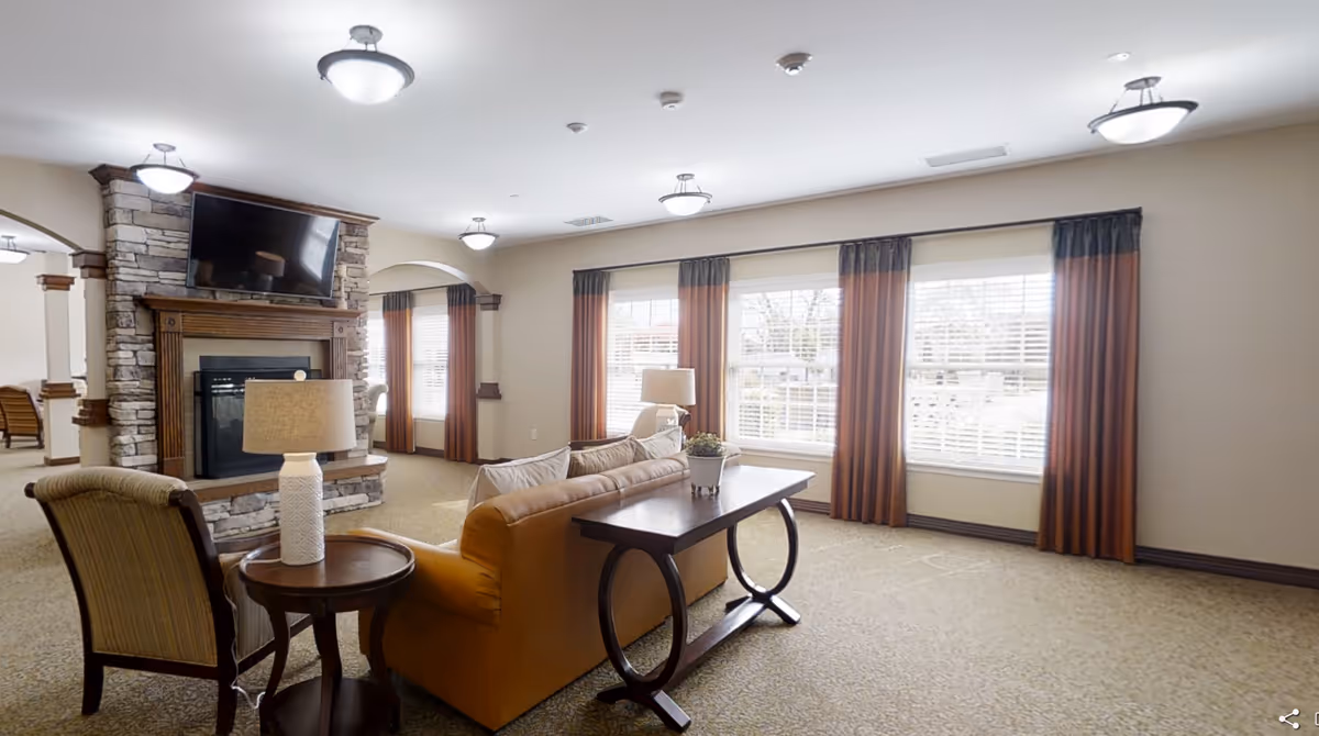 A bright communal living room with sofas and chairs facing a stone fireplace and TV, and large windows with drapes.