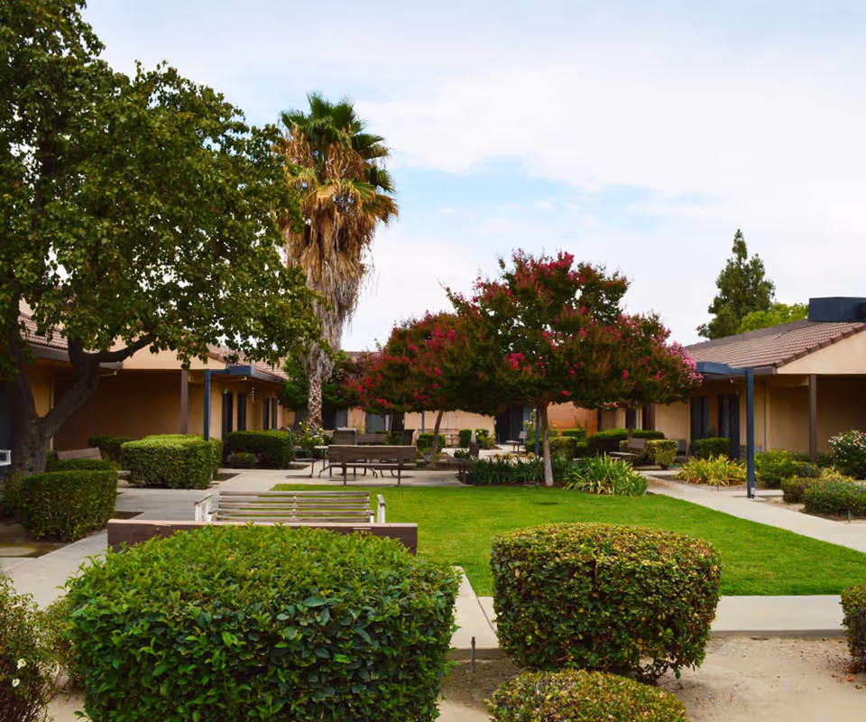 Outdoor courtyard area at Park Visalia Assisted Living & Memory Care featuring green grass, trimmed bushes, trees including a palm tree and flowering tree, benches, and a paved walkway surrounded by single-story buildings with brown roofs.