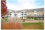 Exterior view of a multi-story senior housing building with balconies, a gazebo, and landscaped grounds including tall grasses and a tree with red leaves.