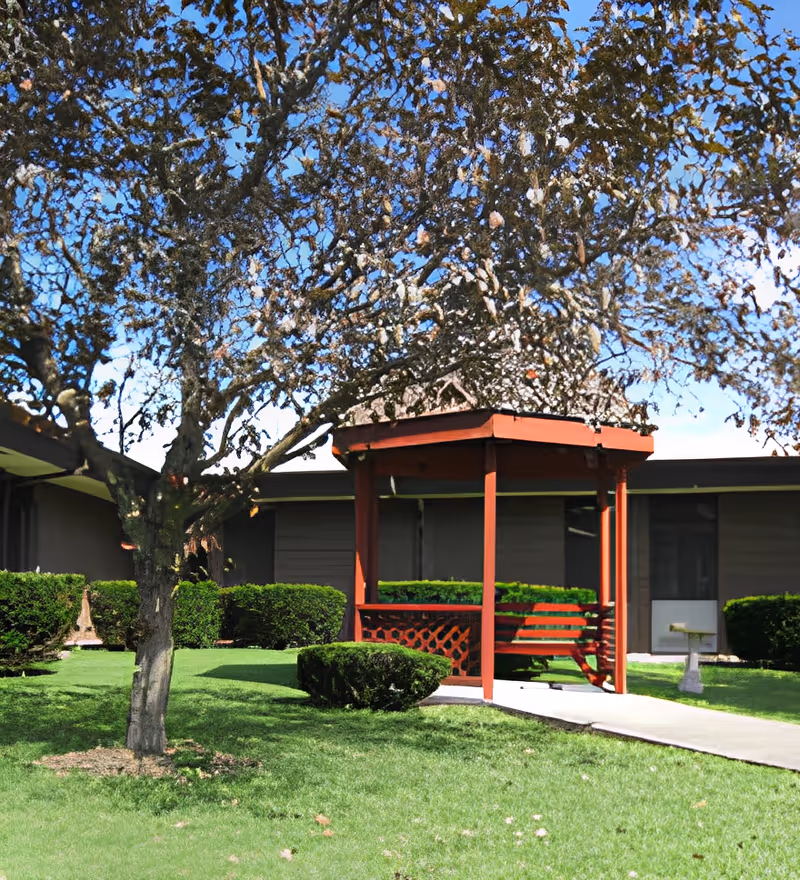 A small red gazebo with a bench in a grassy courtyard under a tree in front of a single-story building.