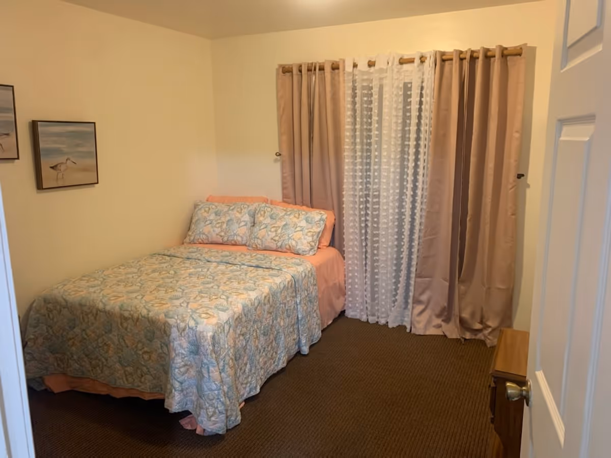 A small bedroom with a bed covered in a floral patterned quilt and matching pillows. The bed has a peach-colored sheet and is positioned against a cream-colored wall. There are two framed pictures of birds on the wall to the left. A window with sheer white curtains and beige drapes is on the wall behind the bed. A wooden nightstand is partially visible on the right side of the image.