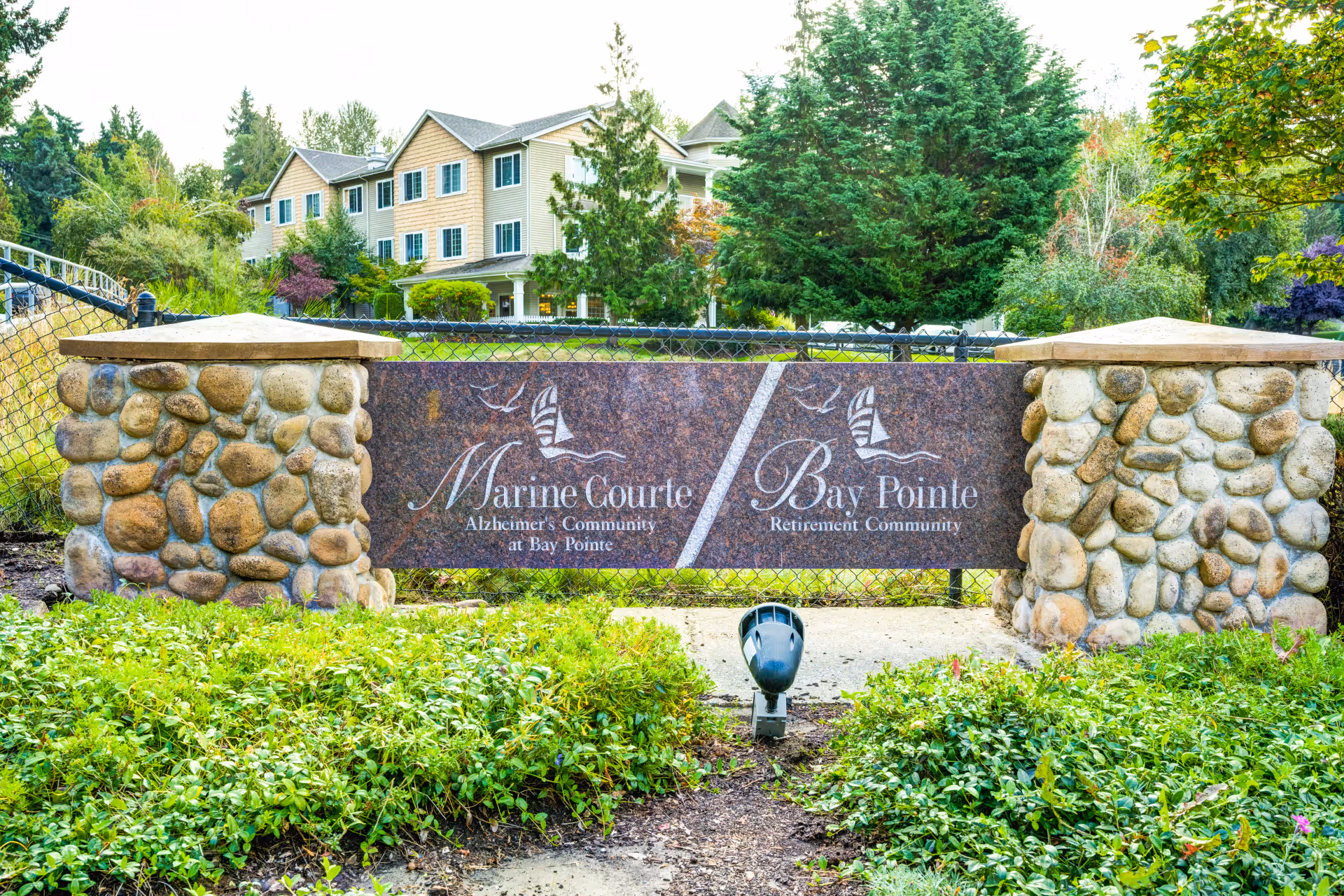 Stone pillars supporting a sign for Marine Courte Alzheimer's Community and Bay Pointe Retirement Community, with a multi-story residential building and trees in the background.