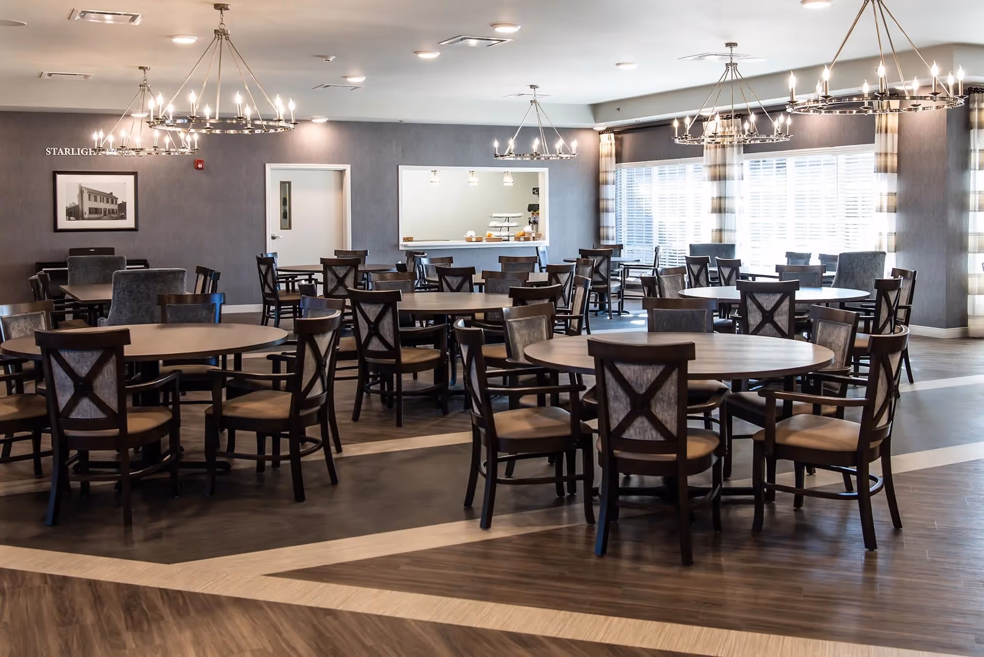 A spacious dining room with multiple round and rectangular wooden tables surrounded by dark wooden chairs with cushioned seats. The room features large windows with striped curtains allowing natural light to fill the space. Modern chandeliers with multiple lights hang from the ceiling. A serving window is visible on the far wall, along with a framed black and white photograph and a door.