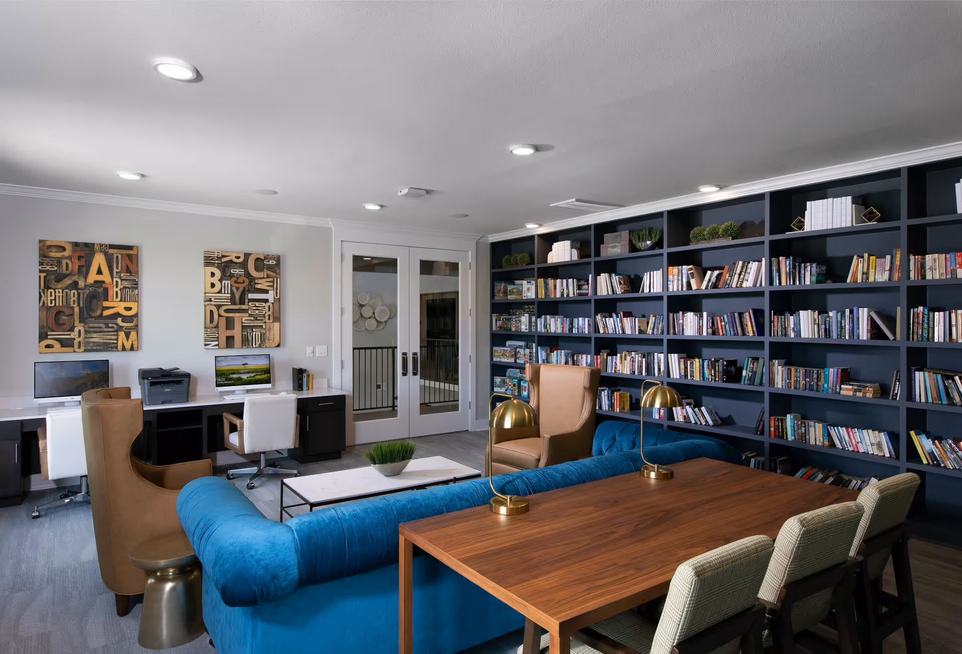 A cozy library and workspace area featuring a large dark blue bookshelf filled with books, a blue velvet sofa, a wooden table with four chairs, two brown armchairs, and a workspace desk with two computers and a printer. The room has modern recessed lighting and decorative wall art with letters.