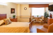 A well-lit bedroom in a senior living facility featuring a bed with a yellow quilt and decorative pillows, a wooden nightstand with a plant, a wall clock, a window with curtains, a small round table with two chairs, a potted plant, a television mounted on the wall, and an armchair.