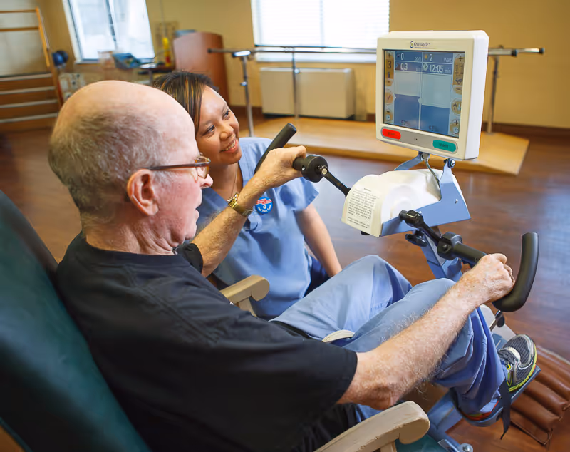 An elderly man using a seated exercise machine with handles and a digital screen, assisted by a smiling female caregiver in blue scrubs in a room with wooden floors and exercise equipment in the background.