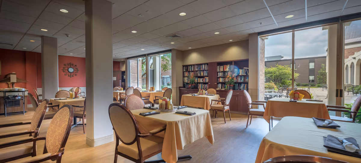 A bright dining room in a senior living facility with tables covered in beige tablecloths, each set with napkins and condiments. The room features large windows overlooking an outdoor patio area with greenery and seating. There is a bookshelf filled with books against one wall and a decorative clock on a red accent wall.