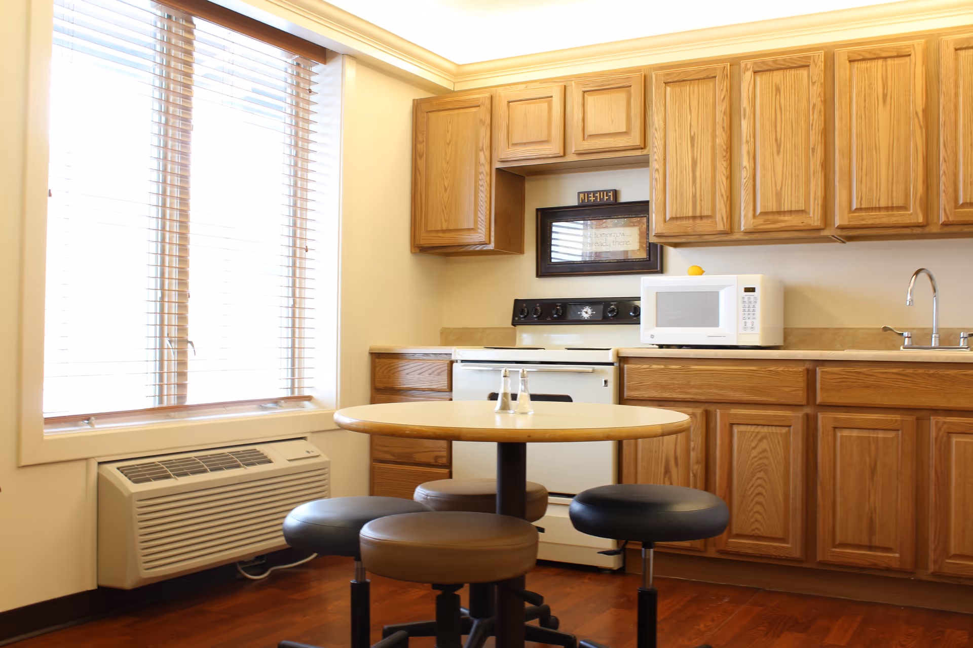 Bright kitchenette with oak cabinets, a stove and microwave, a round table with stools, and a large window with blinds.