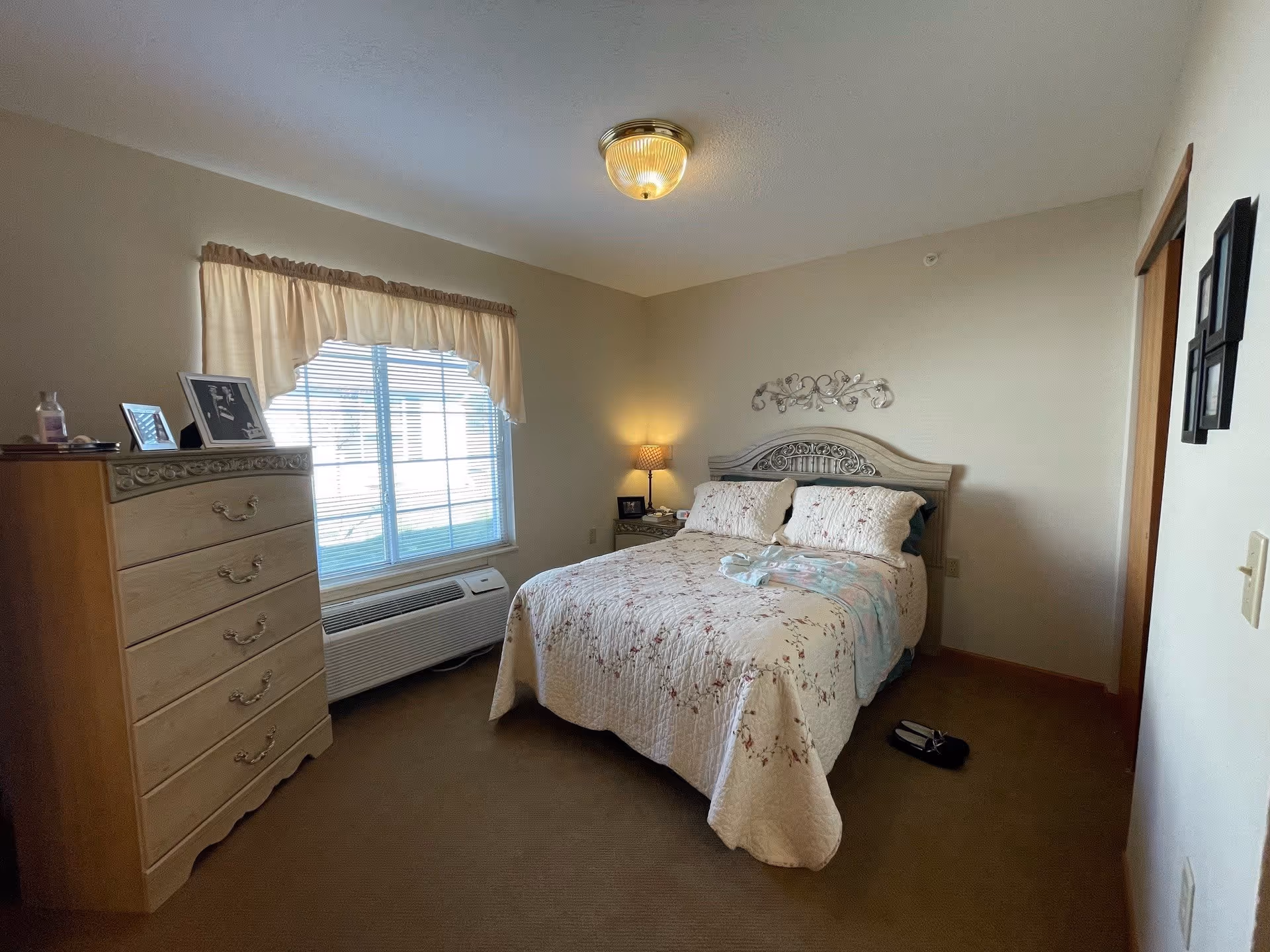 A cozy bedroom with a double bed covered in a floral quilt and pillows. There is a nightstand with a lamp and picture frame next to the bed. A wooden dresser with framed photos and a bottle is positioned near a window with blinds and a valance. The room has beige walls, carpeted floor, and a ceiling light fixture.