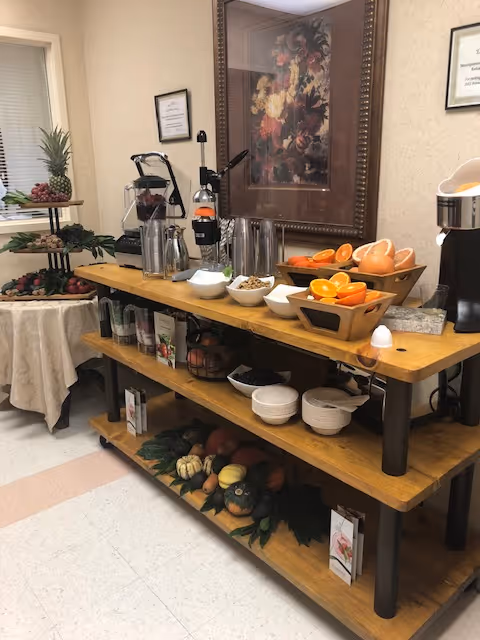 A wooden buffet table with various bowls of fresh fruit including oranges and grapefruit, a juicer machine, and several metal pitchers. There is a tiered stand with grapes and other fruits on a round table covered with a beige tablecloth to the left. The background features a framed floral painting and a beige wall with a small framed sign.