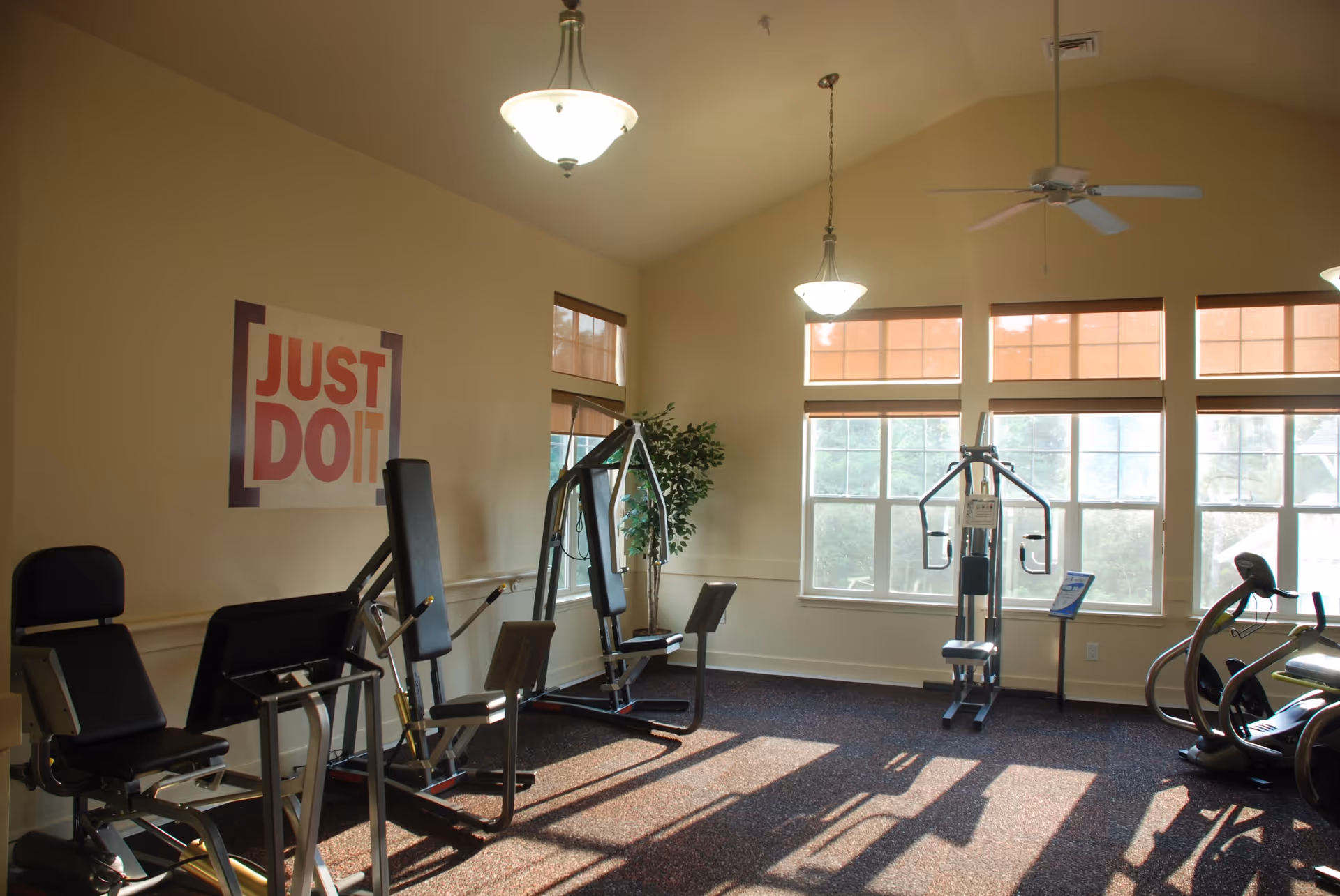 A well-lit exercise room with several pieces of gym equipment including weight machines and an elliptical trainer. Large windows allow natural light to fill the room, and a motivational poster on the wall reads 'JUST DO IT'. The room has beige walls, carpeted flooring, ceiling lights, and a ceiling fan.