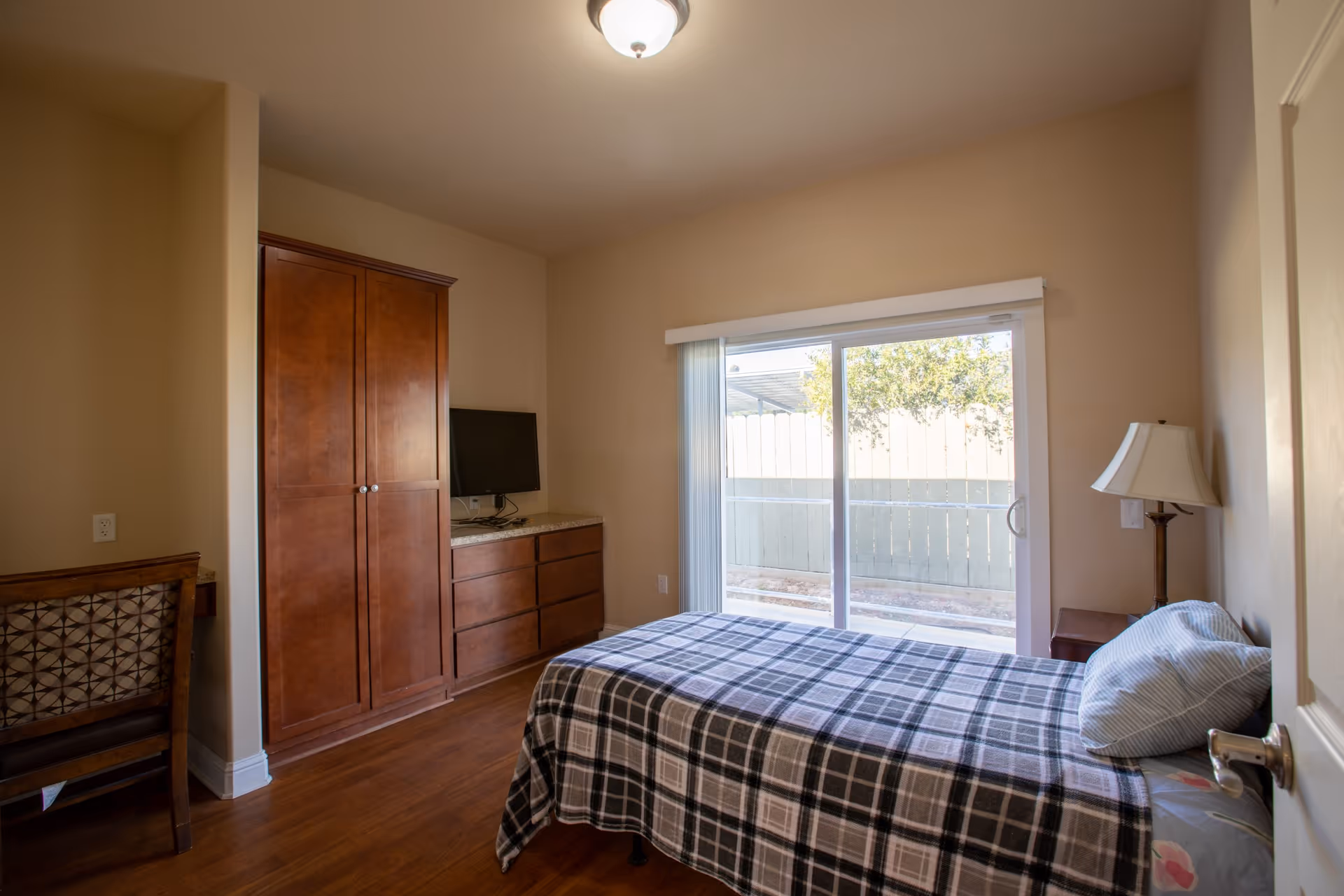 A small bedroom with a single bed covered in a plaid blanket, a pillow, a wooden dresser with a TV on top, a wooden wardrobe, a chair with a patterned backrest, a bedside table with a lamp, and a sliding glass door leading to an outdoor area with a fence and some greenery.