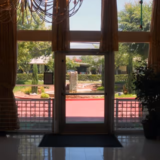View through a glass door from inside a building looking out onto a landscaped outdoor area with trees, bushes, and a stone pillar. The door is framed by large windows and curtains, with a chandelier partially visible at the top left and a potted plant on the right side.