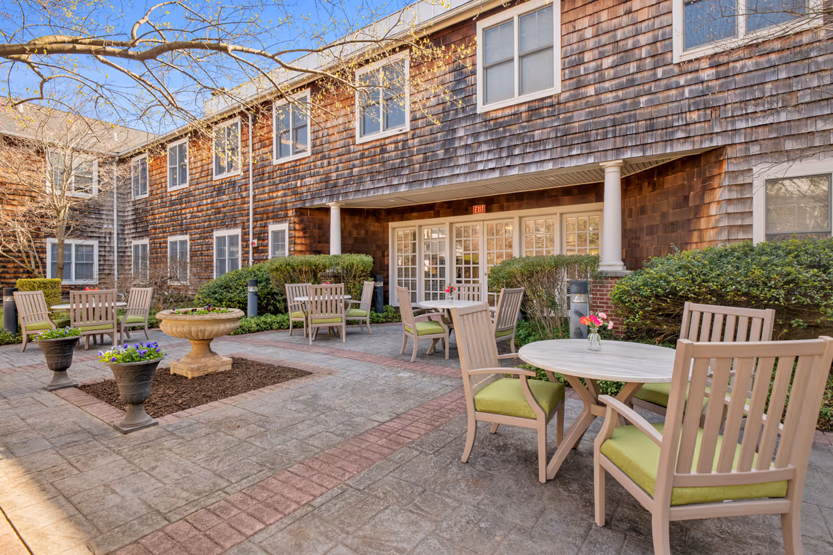 Outdoor courtyard area of a senior living facility with multiple round tables and chairs with green cushions, potted flowers, and a stone birdbath. The building has wood shingle siding and multiple windows, with a covered entrance supported by white columns.
