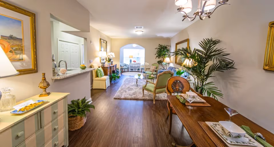 A well-lit, spacious interior of a senior living facility showing a dining area with a wooden table set for two on the right, and a living room area with chairs, a sofa, plants, and lamps in the background. The floor is wooden, and the walls are decorated with framed artwork and mirrors.