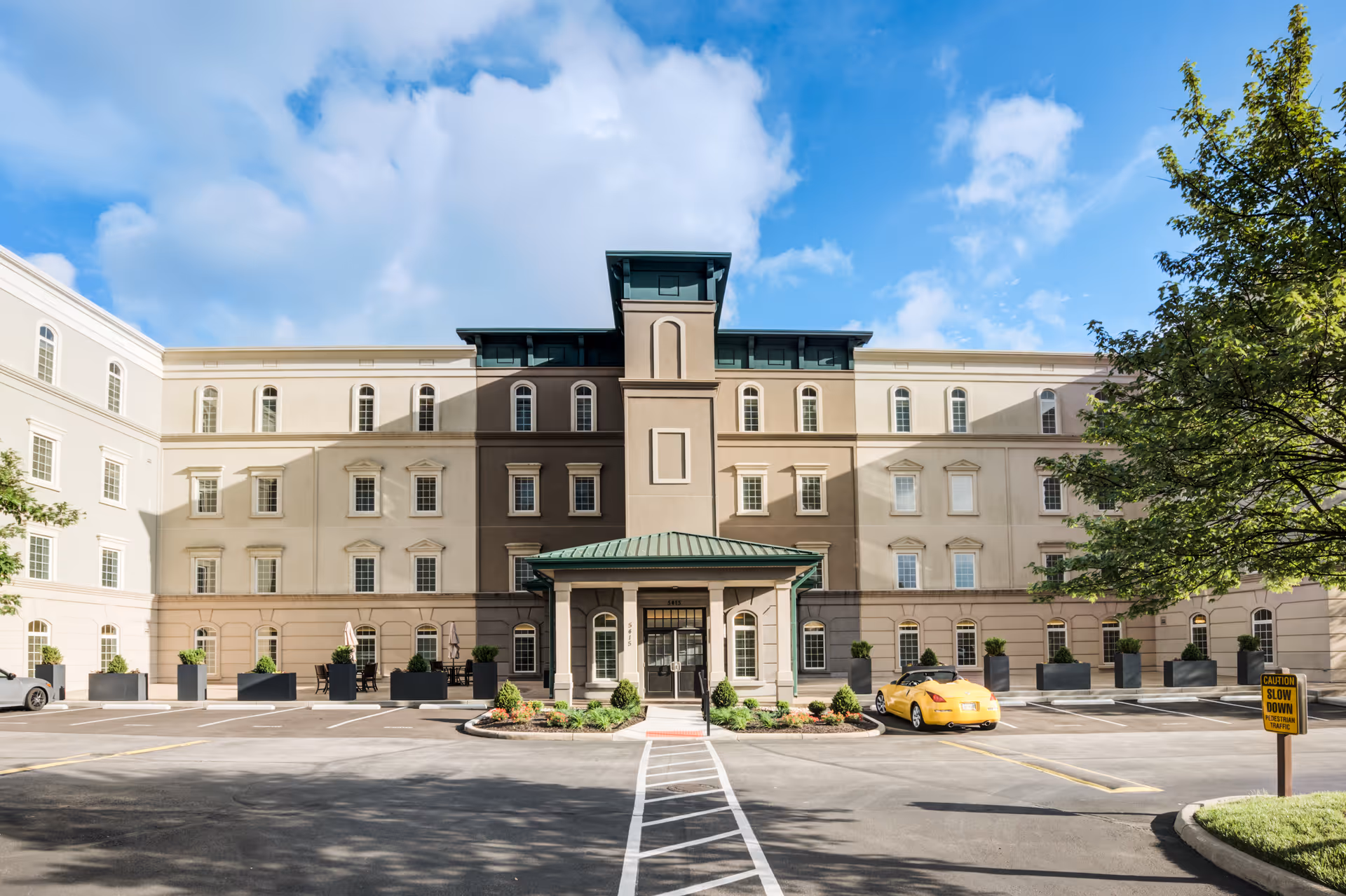 Front exterior view of The Kenwood by Senior Star, a multi-story senior living facility with a central entrance covered by a green roof. The building is beige with multiple windows, and there is a parking lot in front with a yellow car parked. Trees and a blue sky with clouds are visible.