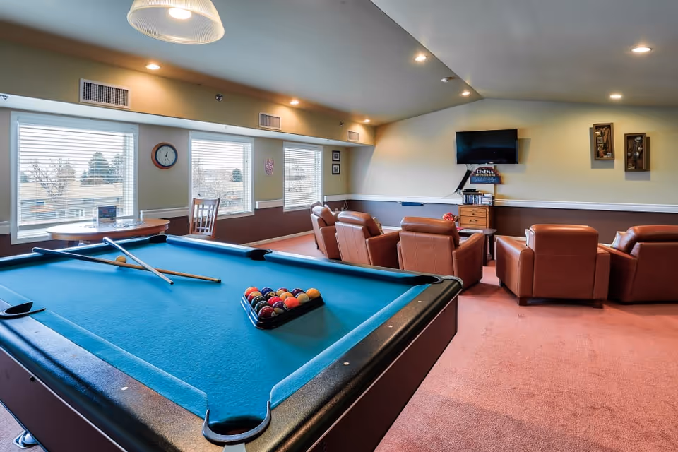 Bright recreation room with a billiards table in the foreground, leather recliners facing a wall-mounted TV, and windows along the left wall.