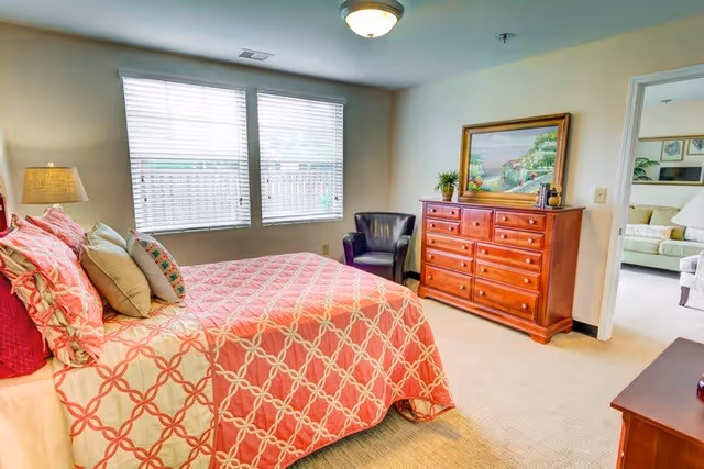 A bright bedroom with a large bed covered in a coral and beige patterned quilt and multiple pillows. There is a wooden dresser with a framed painting and a small plant on top. A black armchair is positioned near the window with white blinds. The room has beige walls and carpet, and an open doorway leads to a living area with a sofa and artwork on the walls.
