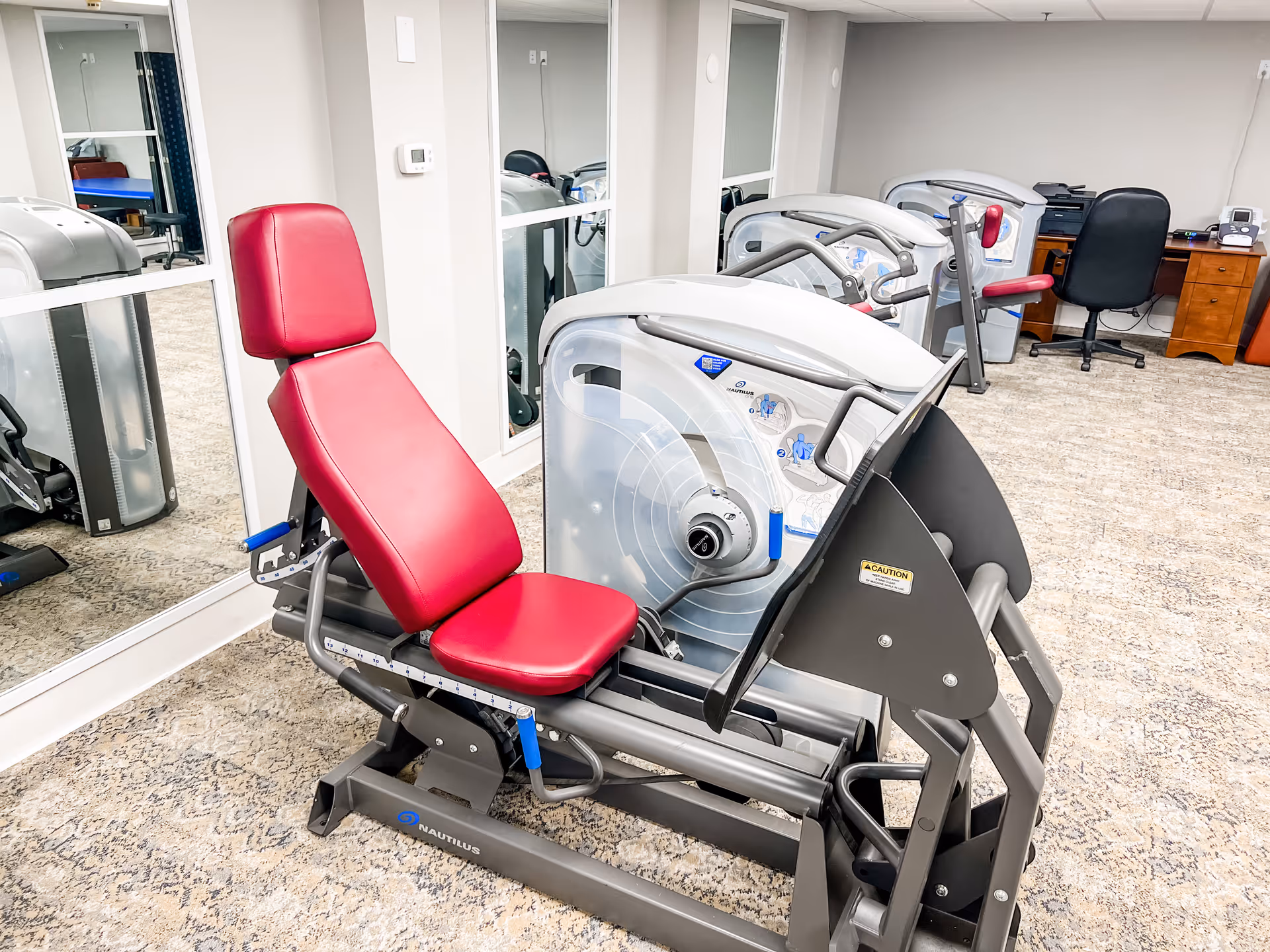 A red-padded Nautilus leg-press exercise machine in a carpeted fitness/therapy room with mirrors and additional equipment.