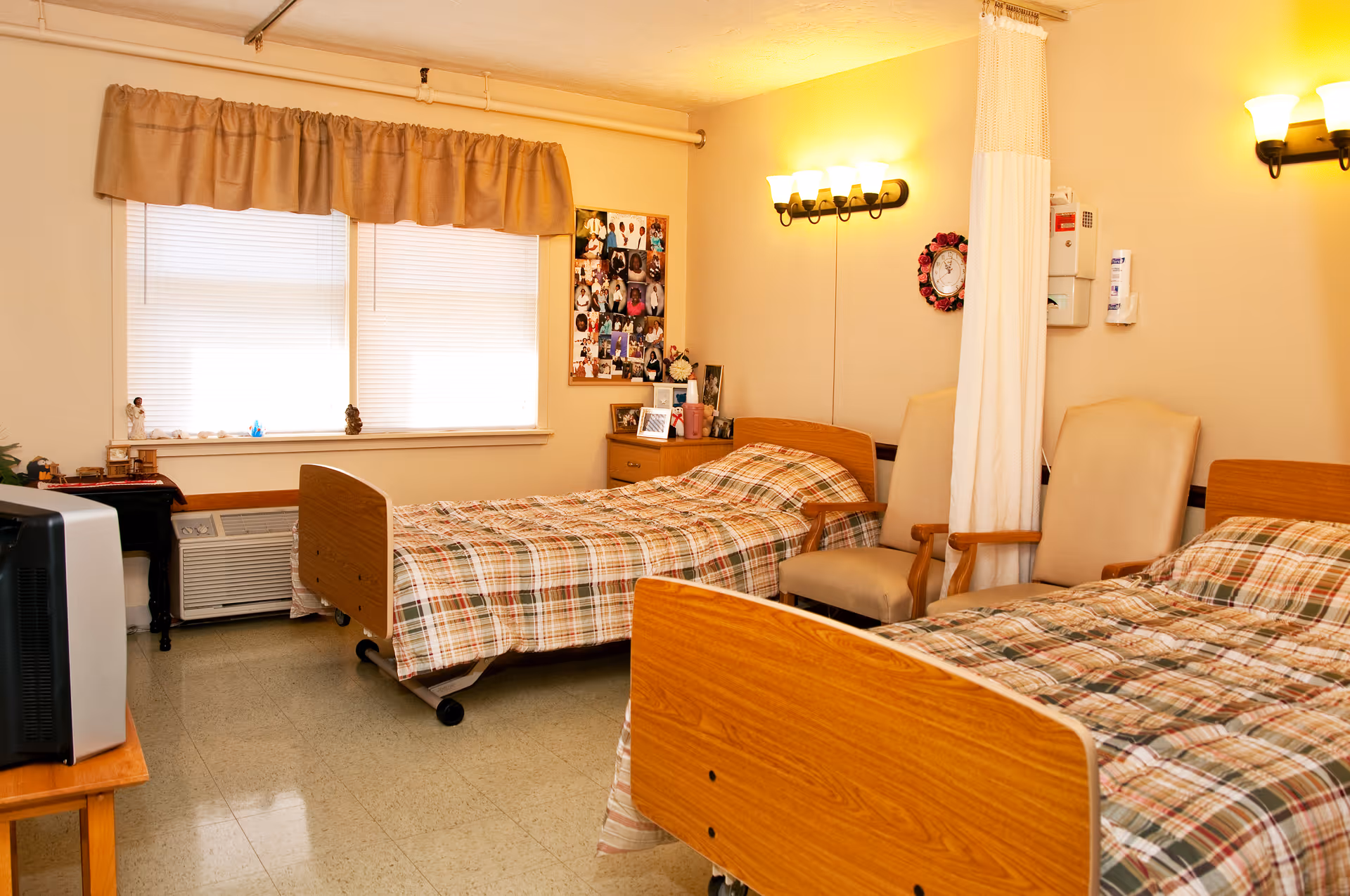 A room with two single beds featuring plaid bedspreads, a window with blinds and a beige valance, two beige armchairs, a small wooden dresser with framed photos and decorations, a TV on a wooden stand, and wall-mounted lights providing warm illumination.