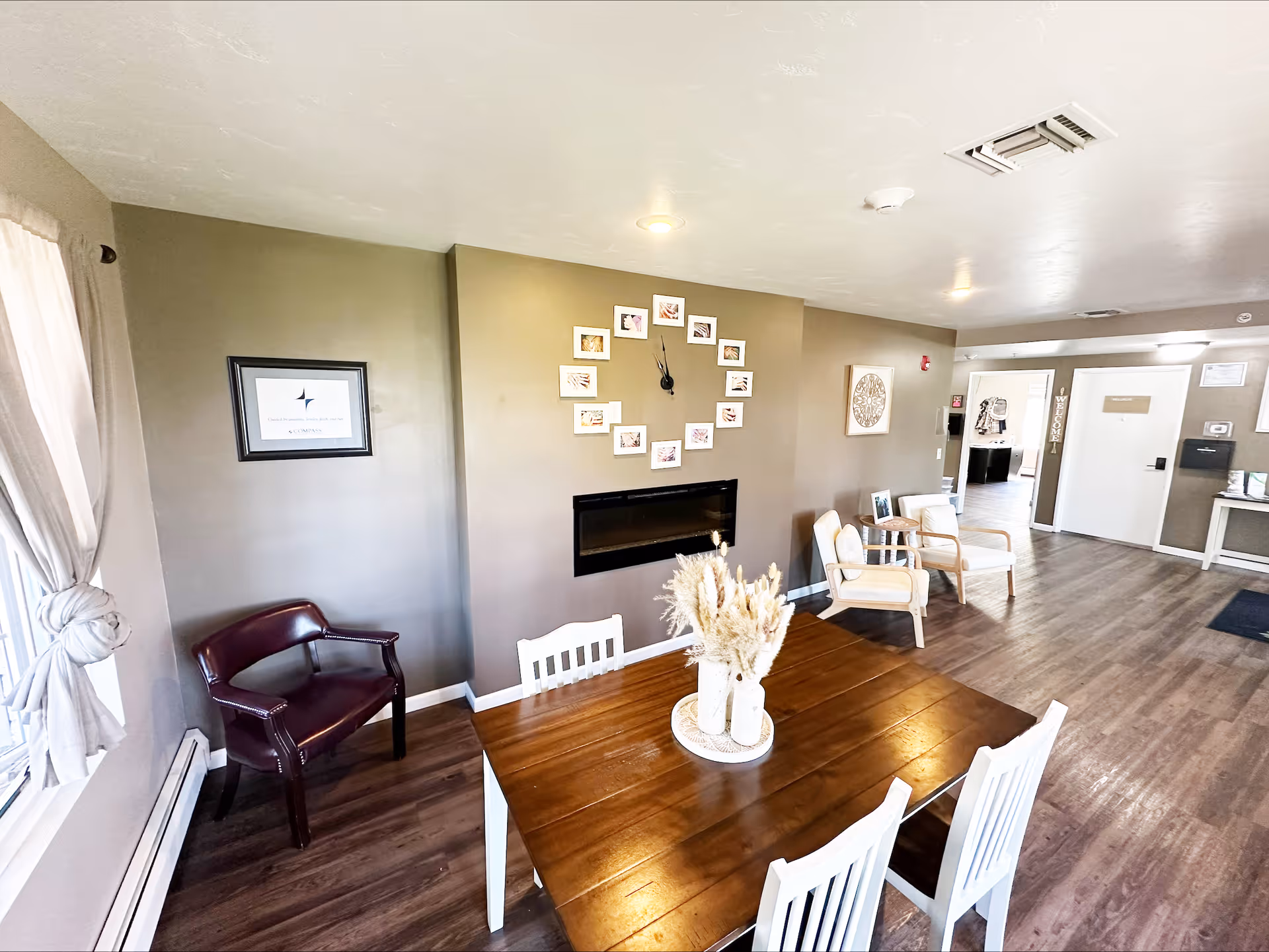 Bright communal interior with a wooden dining table and white chairs, seating area, and a wall-mounted fireplace with framed photos.