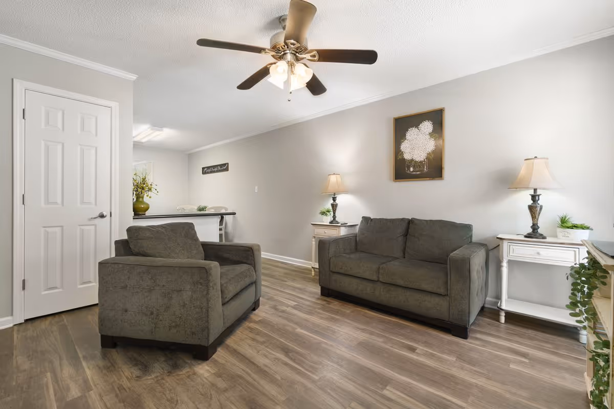 A cozy living room with a gray armchair and a matching gray sofa. There are two white side tables with lamps on either side of the sofa. A framed floral artwork hangs on the wall above the sofa. The room has wood flooring, a ceiling fan with lights, and neutral-colored walls. In the background, a kitchen counter with two chairs and a vase with flowers is visible.