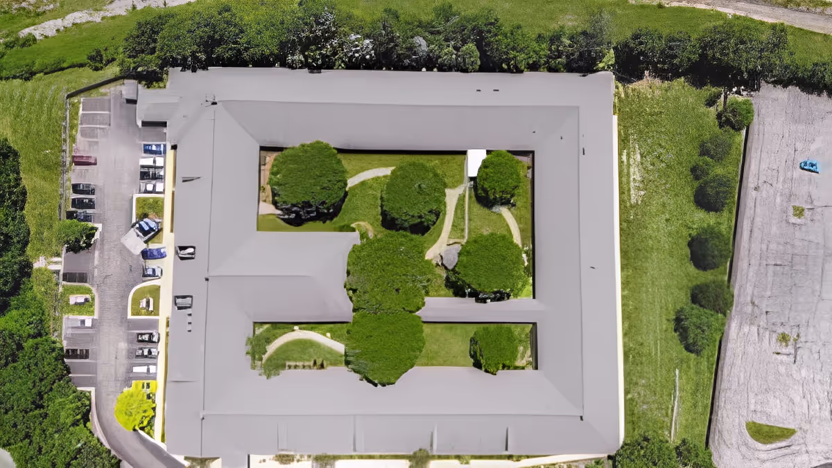 Aerial view of a senior living facility named Rittenhouse Village At Northside showing a large rectangular building with a central courtyard containing trees and walking paths. Surrounding the building are parking areas, greenery, and open spaces.
