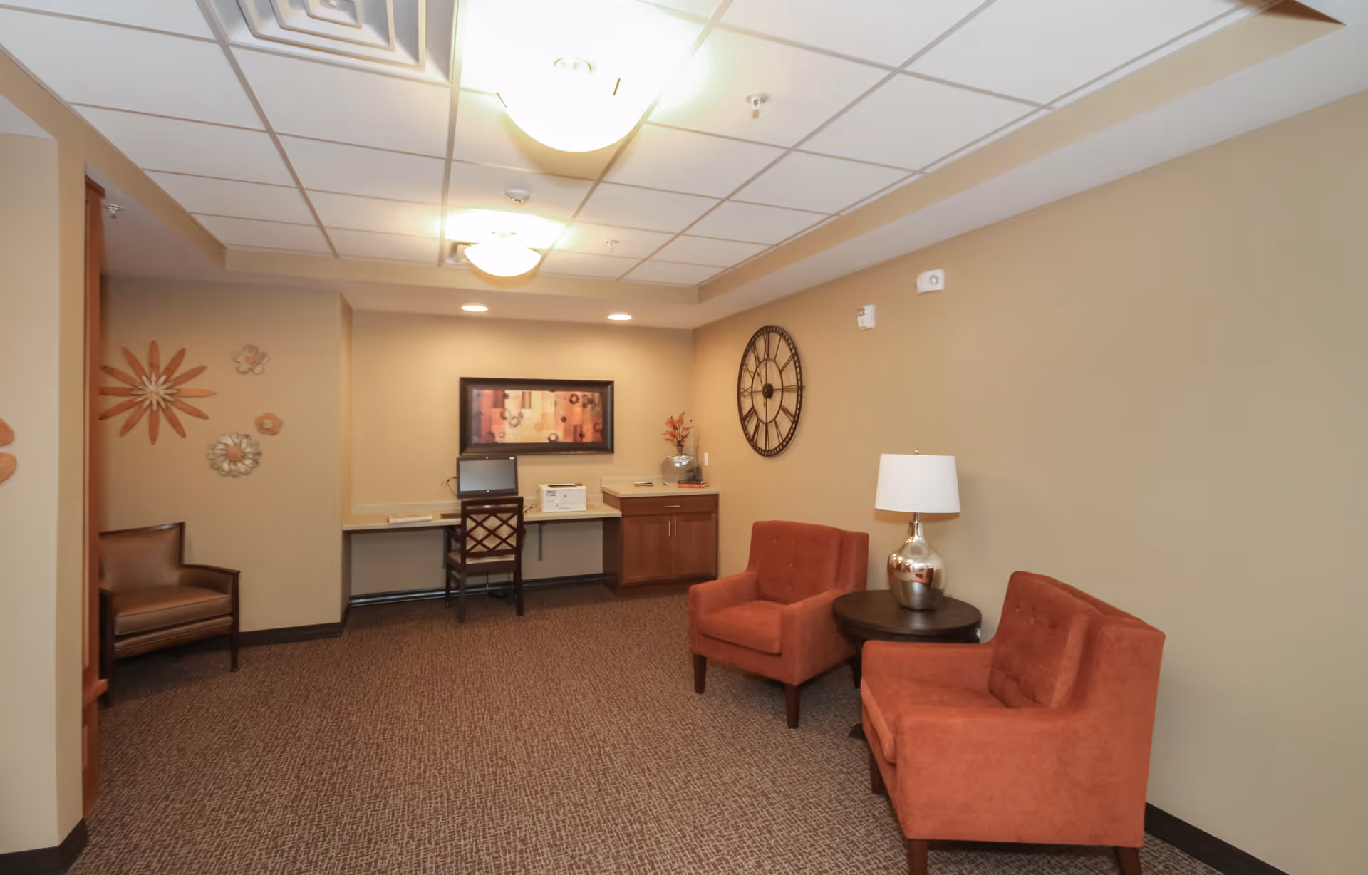 A cozy interior room with beige walls and carpeted floor featuring two orange armchairs with a round wooden side table and a silver lamp between them. On the far wall, there is a large decorative clock and a framed abstract painting above a desk with a chair, computer, and printer. Decorative metal flowers adorn the left wall.