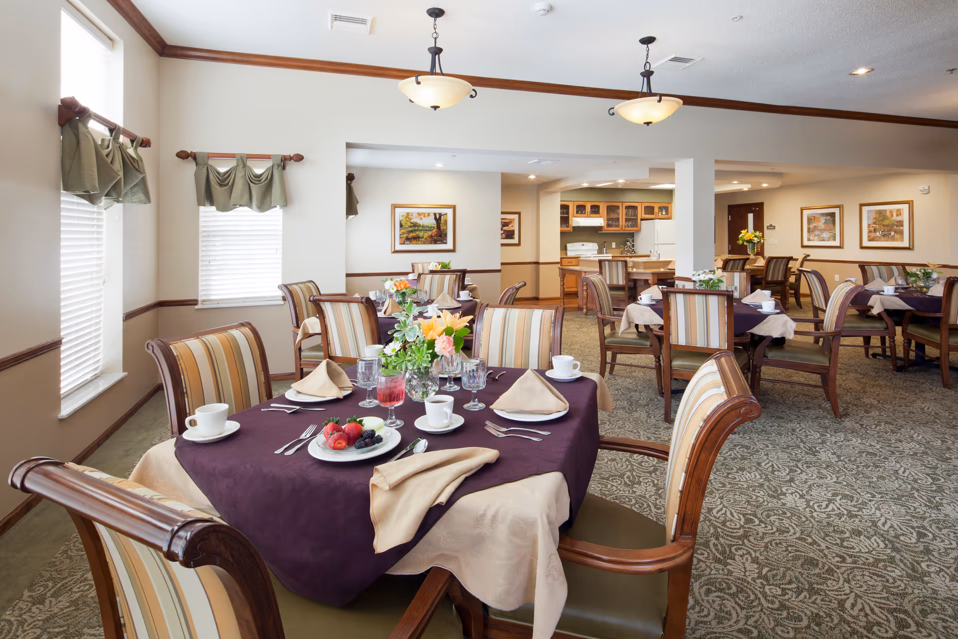 A bright dining room with several tables covered in beige and dark purple tablecloths, each set with cups, glasses, silverware, and napkins. The chairs have wooden frames with striped upholstery. The room has carpeted floors, light walls with framed artwork, and windows with green valances. In the background, there is a kitchen area with wooden cabinets, a stove, and a refrigerator.